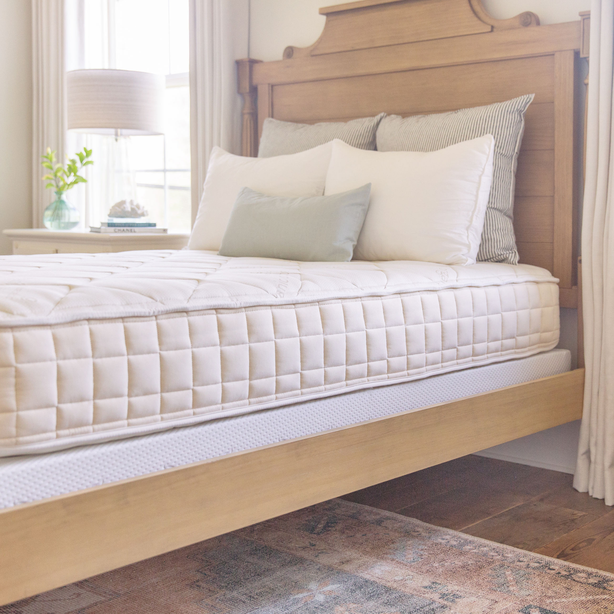 A Naturepedic organic cotton and wool mattress on a light wood platform bed in a sunlit bedroom, with the quilted organic cover and ventilated base edge clearly visible