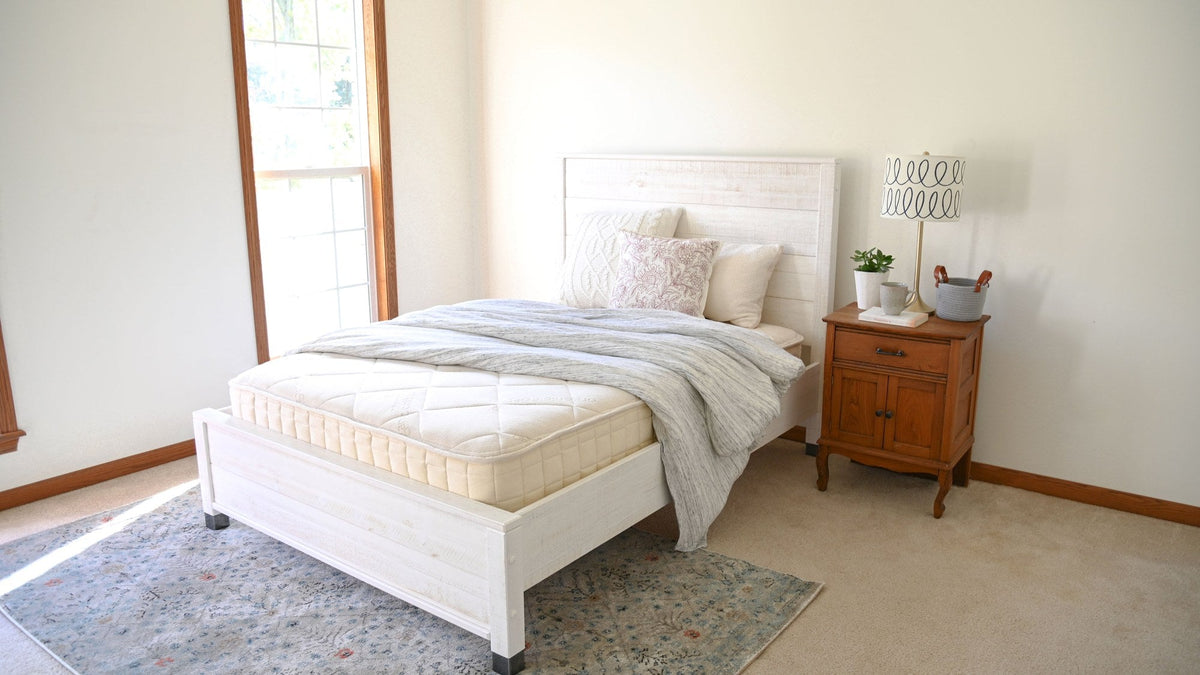 Naturepedic organic mattress on a whitewashed wooden bed frame in a bright, simply furnished spare bedroom with a knit throw, cream and rose pillows, an area rug and a wooden nightstand with a lamp and plant