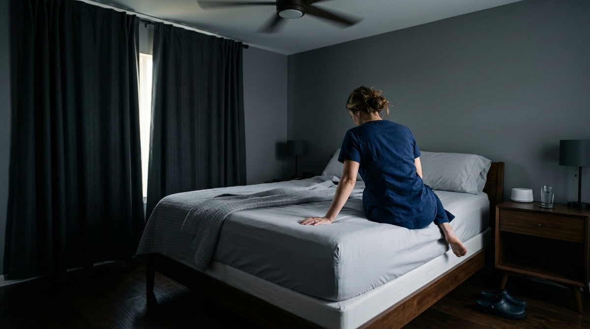 Nurse in scrubs home from a night shift sitting on the edge of a cooling mattress in a darkened bedroom with blackout curtains drawn against midday sunlight