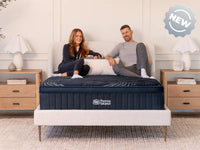 Man and woman sitting on a bed with a mattress, surrounded by a homey bedroom setting.