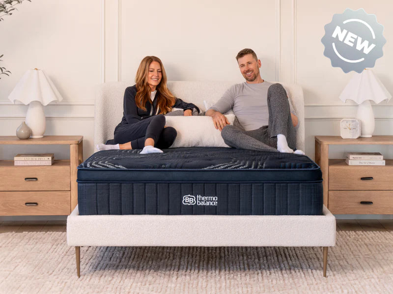 Man and woman sitting on a bed with a mattress, surrounded by a homey bedroom setting.